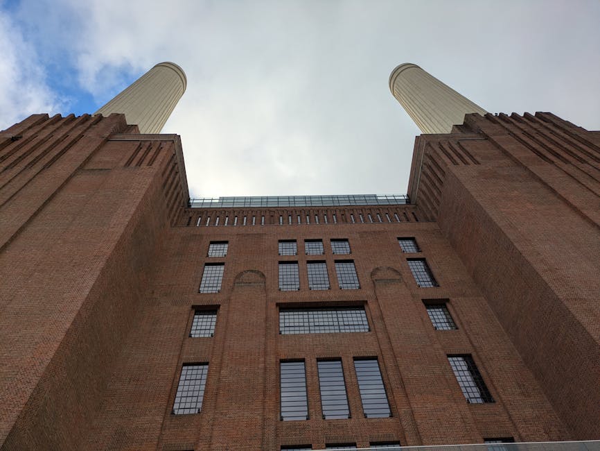 An upward view of Battersea Power Station's brick façade, featuring multiple rectangular windows with black frames, some with reflective glass panes, and two tall, cylindrical chimneys extending into