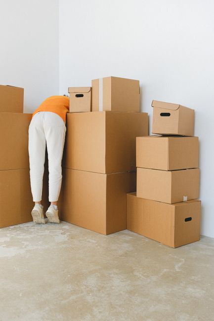 A man and a woman are engaged in a home relocation process outdoors, with the woman placing a cardboard box into the open trunk of a red car, while the man carries a similar box towards a wooden fence